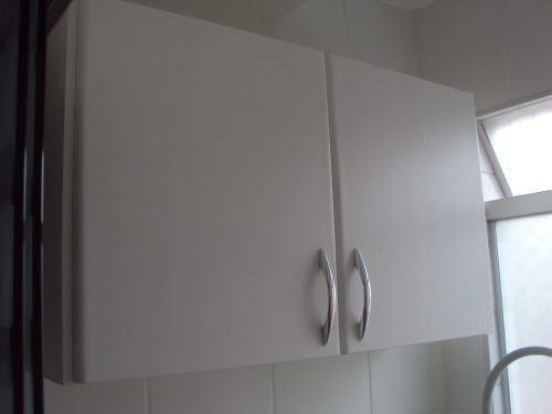 Armário com duas portas em fórmica branca, prateleira interna, puxadores em aço cromado acima do gabinete da cozinha.
